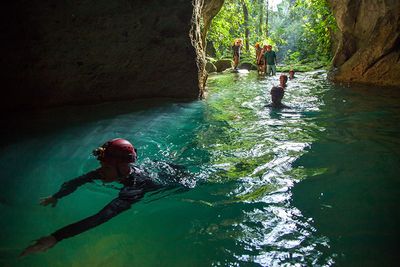 man with red helmet swimming into cave entrance with people behind him